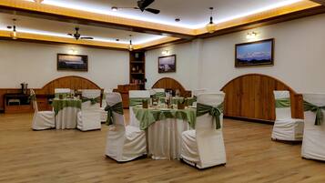 A banquet hall with round tables, white chairs with green sashes, wooden paneling, ceiling fans, and framed pictures on the walls.