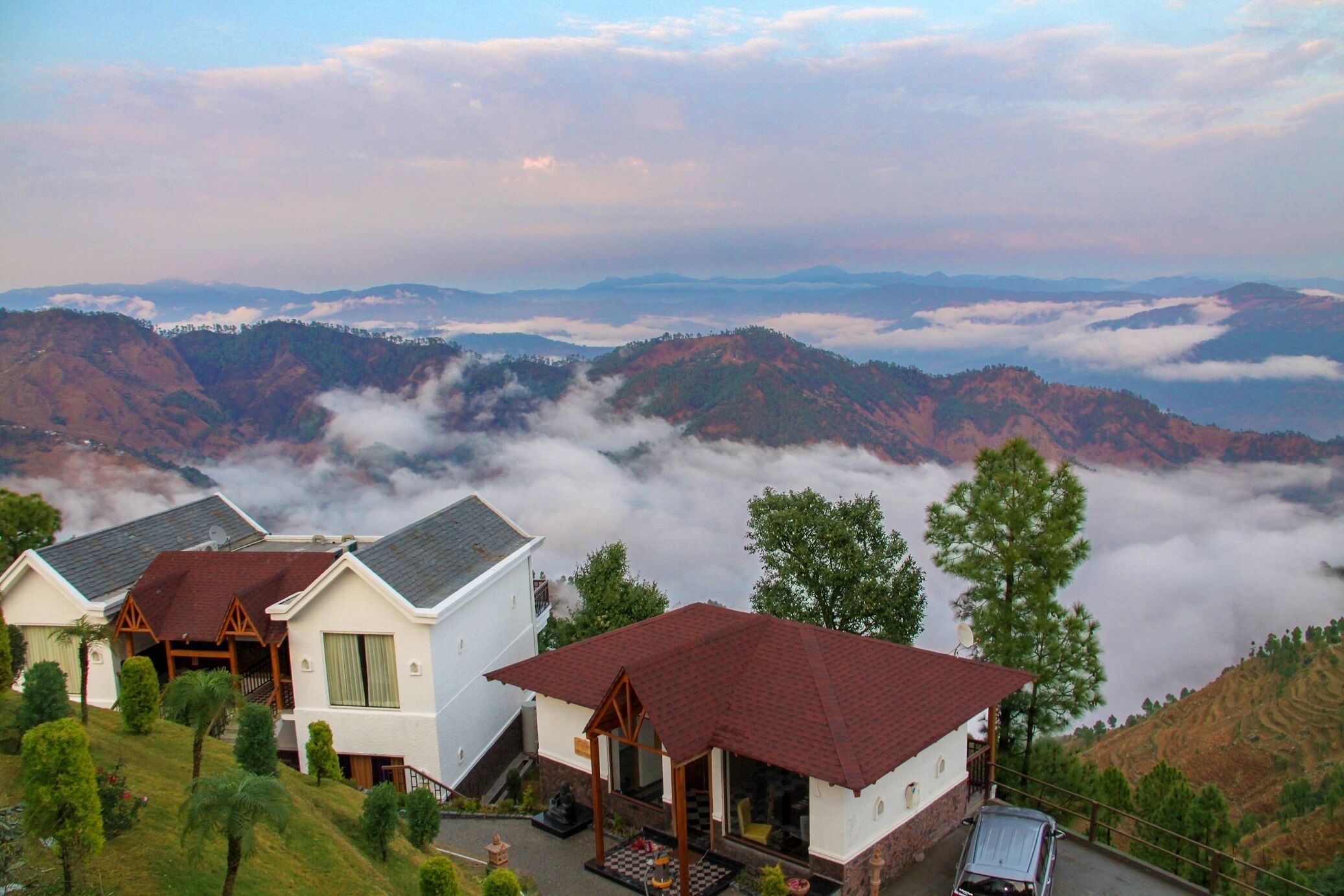 A hillside hotel with multiple buildings, each featuring a distinct architectural style with red roofs and white walls. The surrounding landscape includes rolling hills and a valley below, partially obscured by clouds.