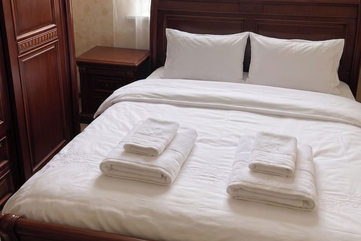 Una cama bien hecha con sábanas blancas y toallas dobladas, un cabecero de madera, una mesita de noche y un armario.