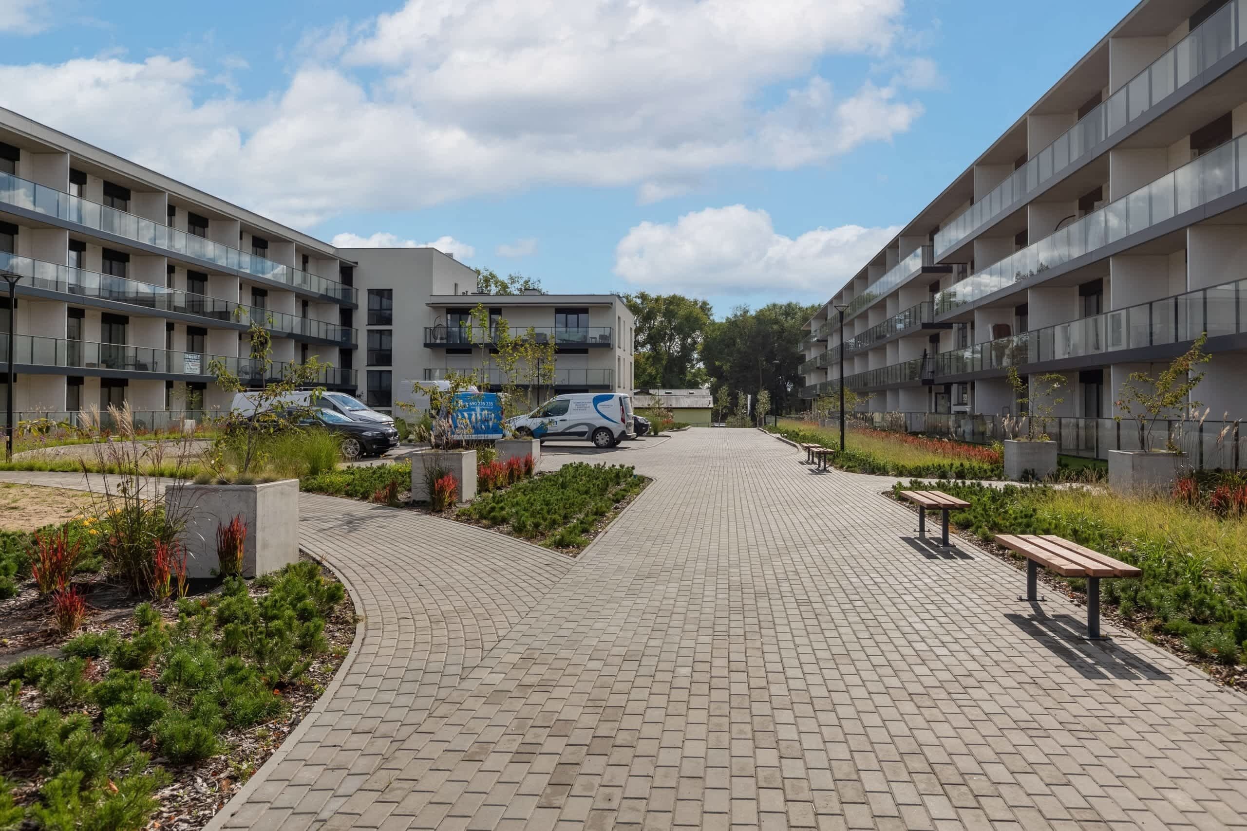 Un moderno complejo de apartamentos con un camino pavimentado, bancos y un coche en el estacionamiento.