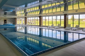 An indoor swimming pool with clear glass walls, lounge chairs, and a view of the outdoors.