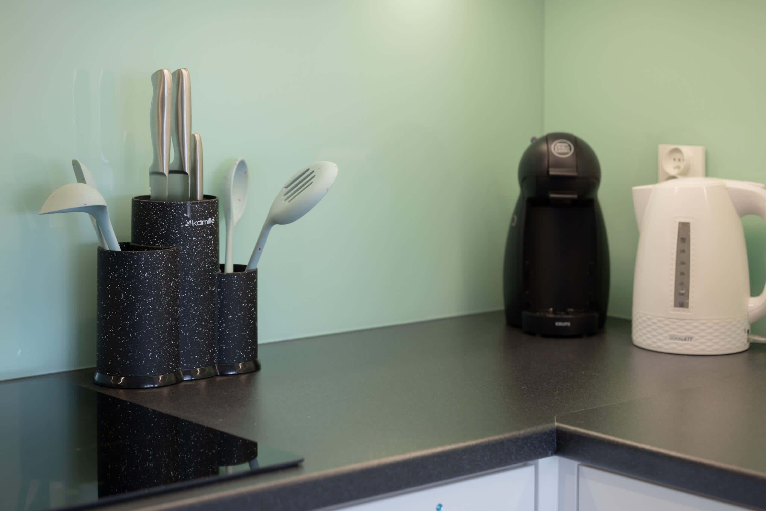 Una encimera de cocina con un recipiente negro con motas que contiene cuchillos y cucharas, una cafetera negra y un hervidor eléctrico blanco.