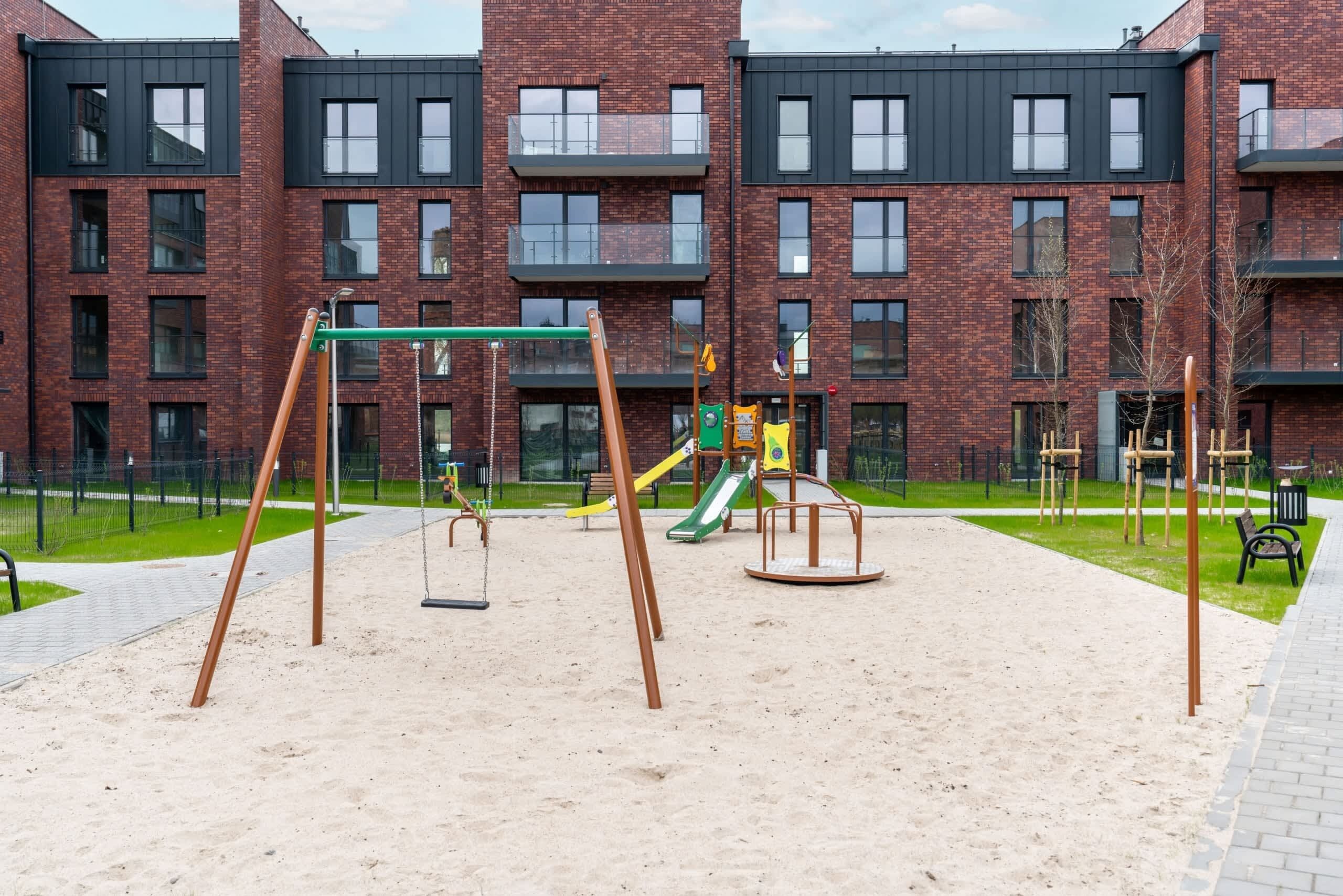 A playground with swings and slides, surrounded by a brick building with multiple windows and balconies.