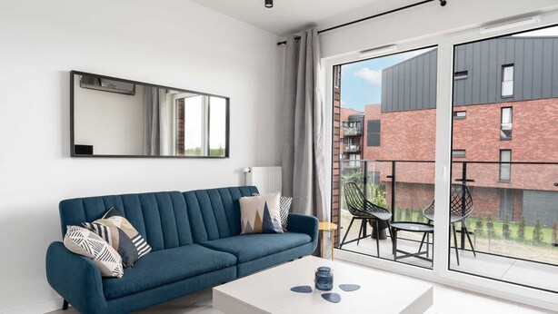 A modern living room with a blue sofa, a white coffee table, and a large window offering a view of a brick building. - Sunny Apartment with Balcony | Parking | Lake View (Poznan)