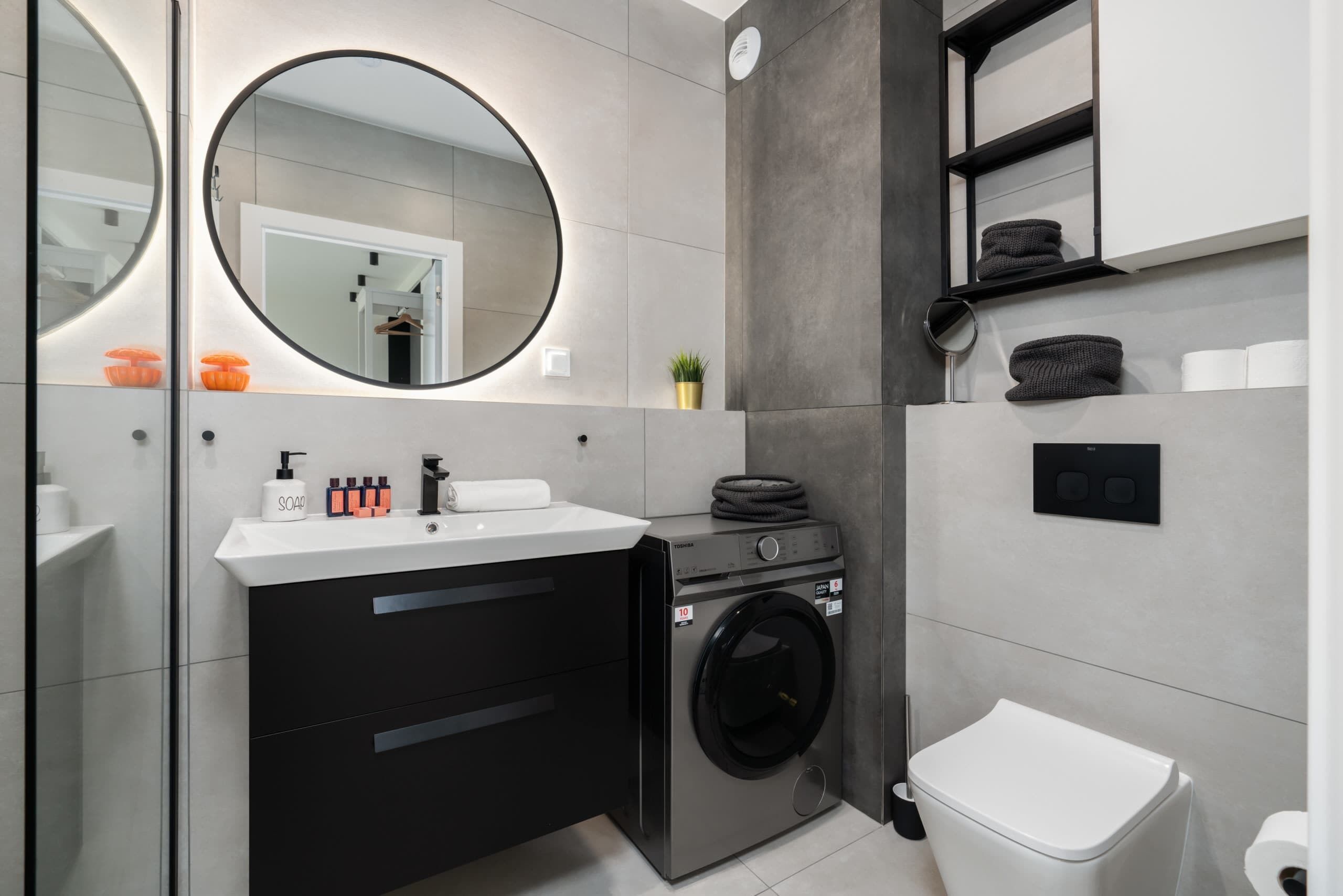 A modern bathroom with a black and white sink, a round mirror, a washing machine, and a white toilet.