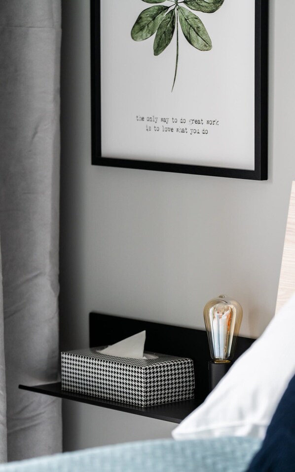 Una cita inspiradora enmarcada en una pared, una mesita de noche con una caja de pañuelos y una lámpara, y una cama con un almohadón blanco.