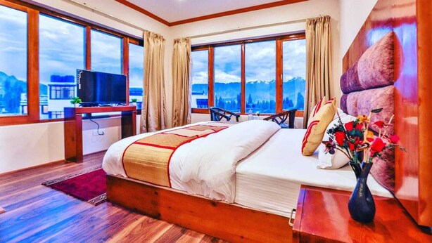 A hotel room with a large bed, a desk with a TV, and a view of mountains through large windows.