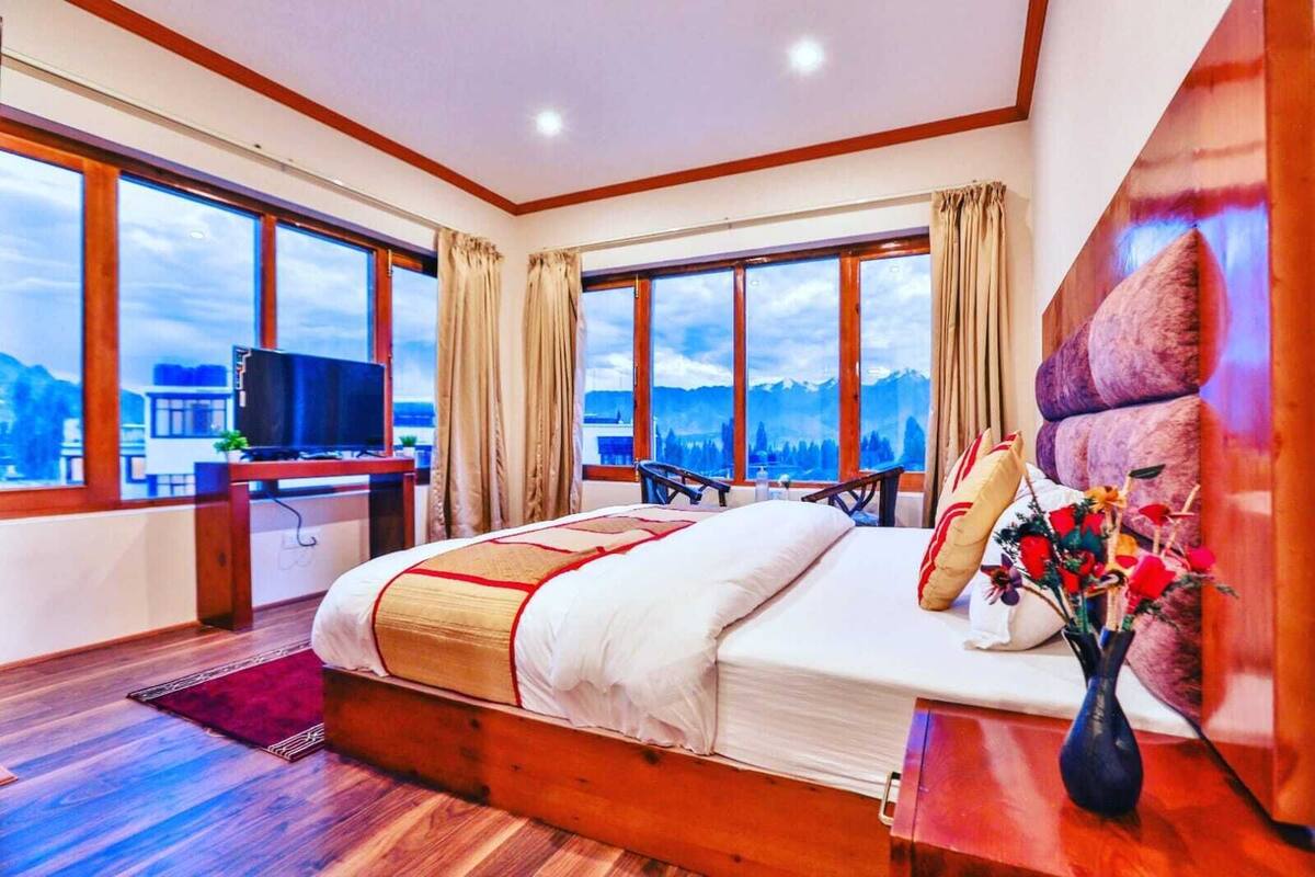 A hotel room with a large bed, a desk with a TV, and a view of mountains through large windows.
