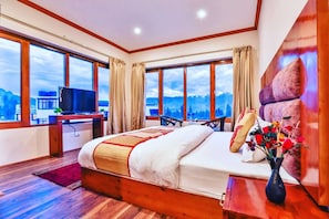 A hotel room with a large bed, a desk with a TV, and a view of mountains through large windows. - De Solace Hotel Leh (Leh)
