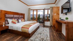 A bedroom with a large bed, wooden headboard, a desk, a TV, and a balcony with a mountain view.