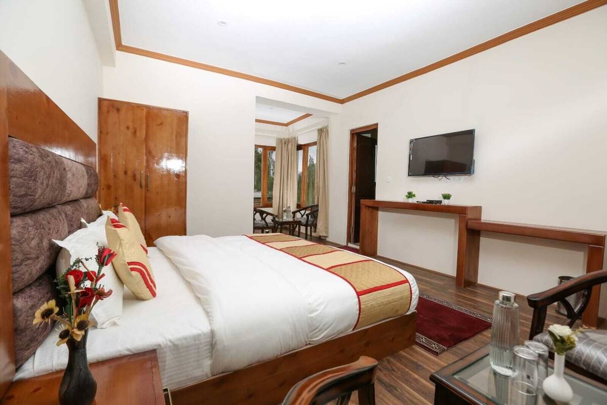 A hotel room with a wooden bed, a TV mounted on the wall, a desk with a chair, and a view of the outside through a window.
