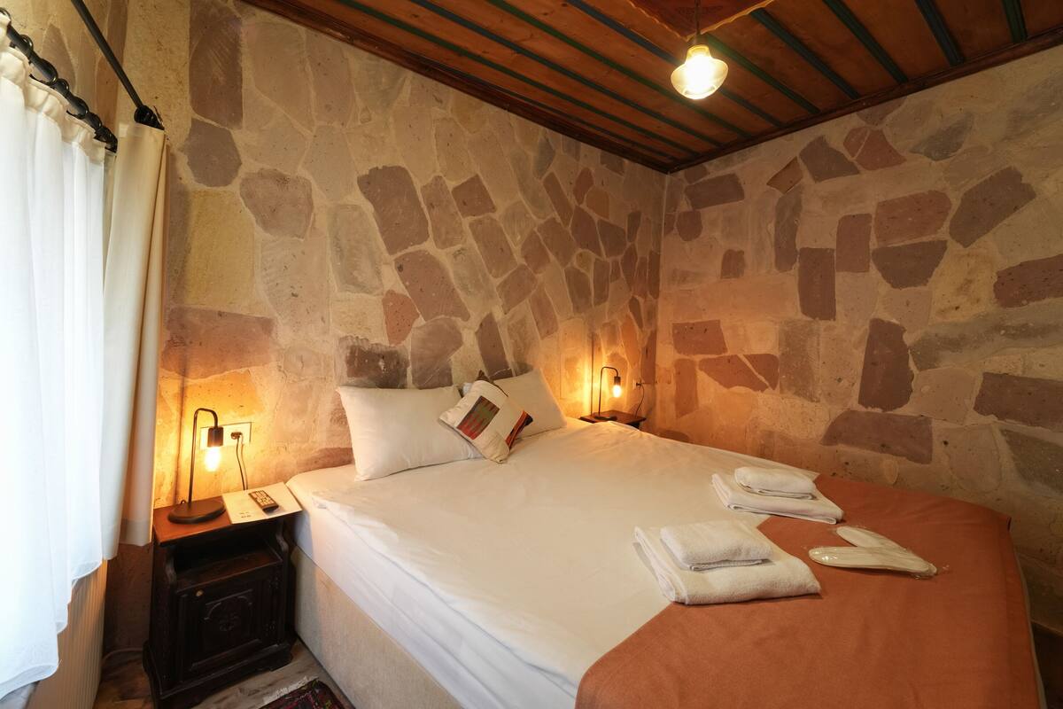 Un dormitorio con pared de piedra, techo de madera, cama con ropa blanca, dos lámparas de noche y una mesita de noche con una lámpara y un libro.