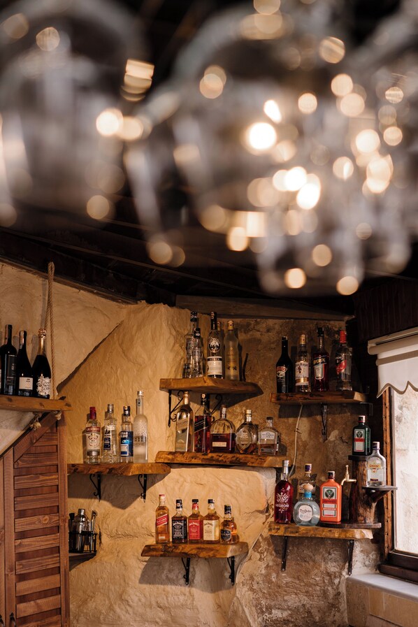 A bar area with shelves of various liquor bottles, wooden shelves, and a rustic stone wall.