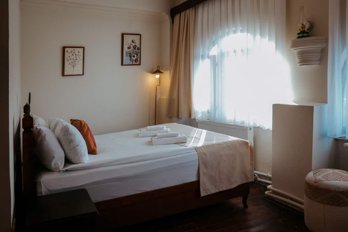 Una cama bien hecha con sábanas blancas y un cabecero marrón, una lámpara de noche y dos impresiones enmarcadas de motivos botánicos en la pared.