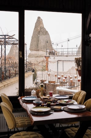 Una mesa de comedor dispuesta con platos y cubiertos, una ventana con vista a una construcción de piedra y una cerca de metal.