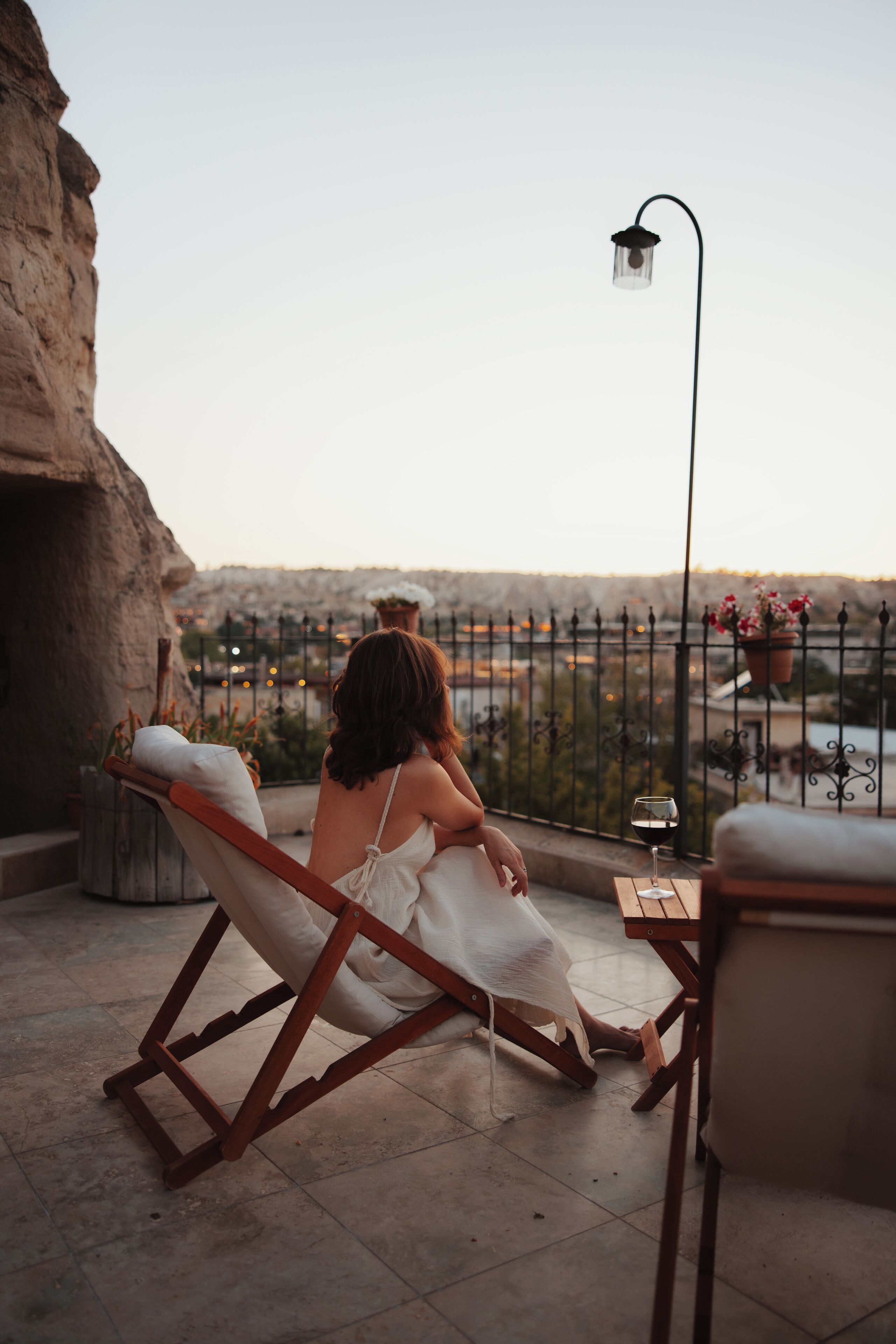A woman sitting on a wooden lounge chair on a balcony overlooking a cityscape.