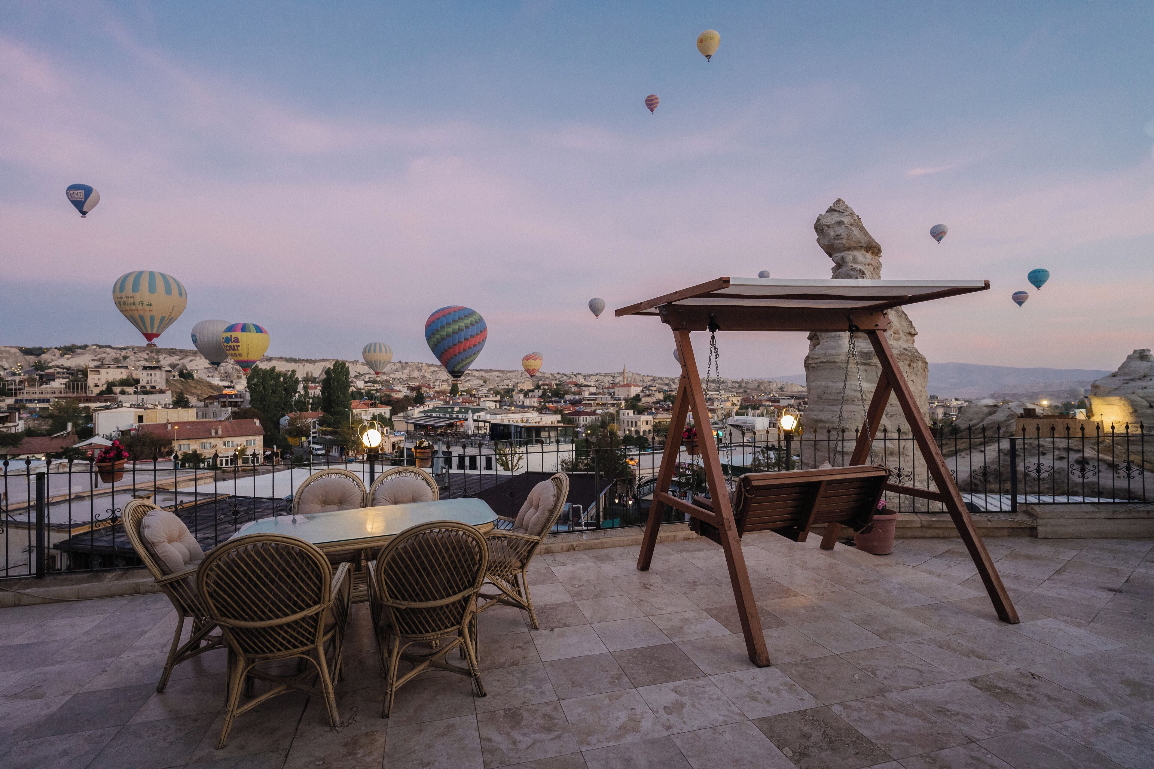 Eine Terrasse mit Korbmöbeln und einer Schaukel mit Blick auf eine Stadtlandschaft, in deren Himmel Heißluftballons schweben.