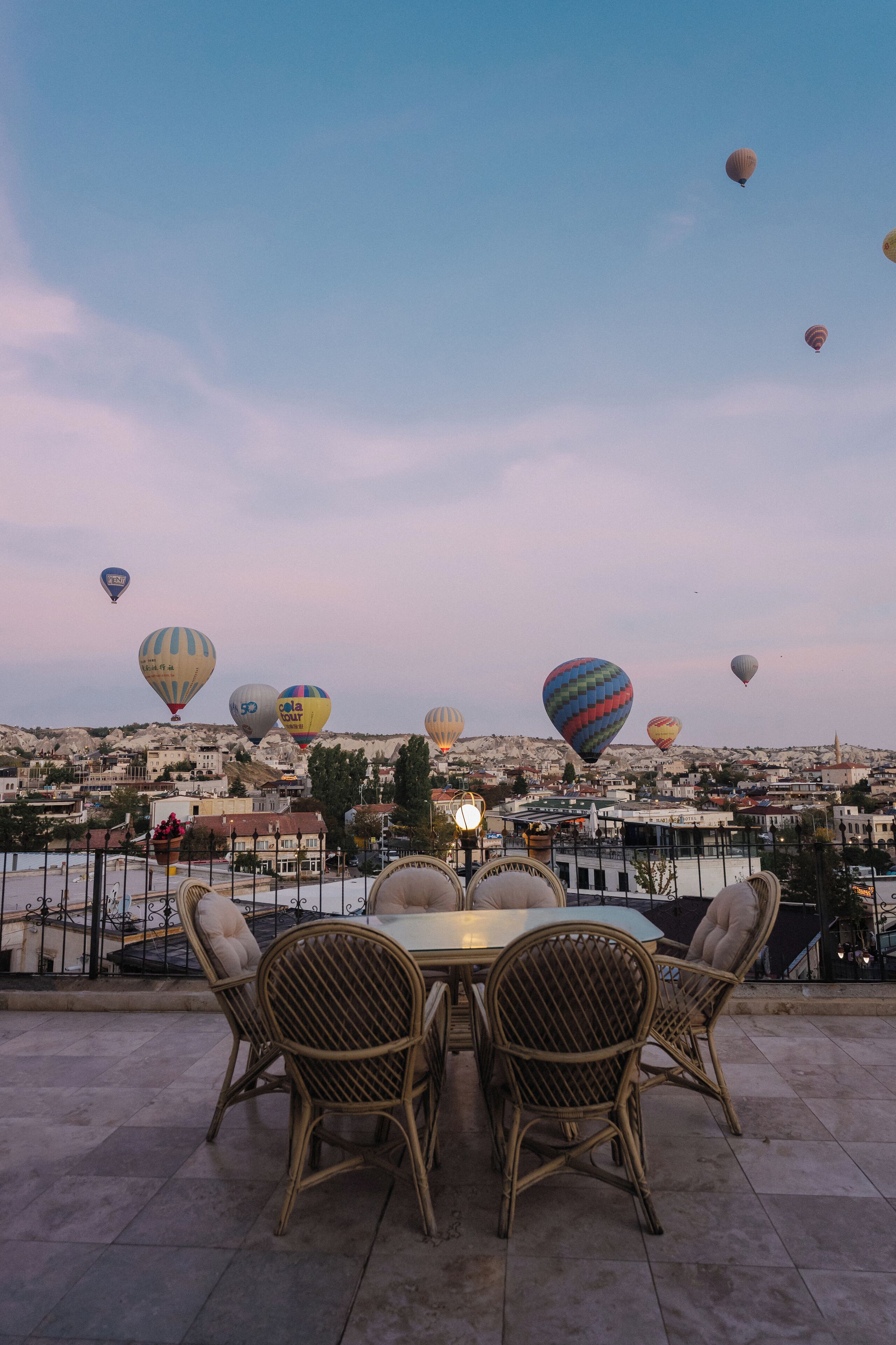 A terrace with wicker chairs and a table, overlooking a cityscape with hot air balloons in the sky.