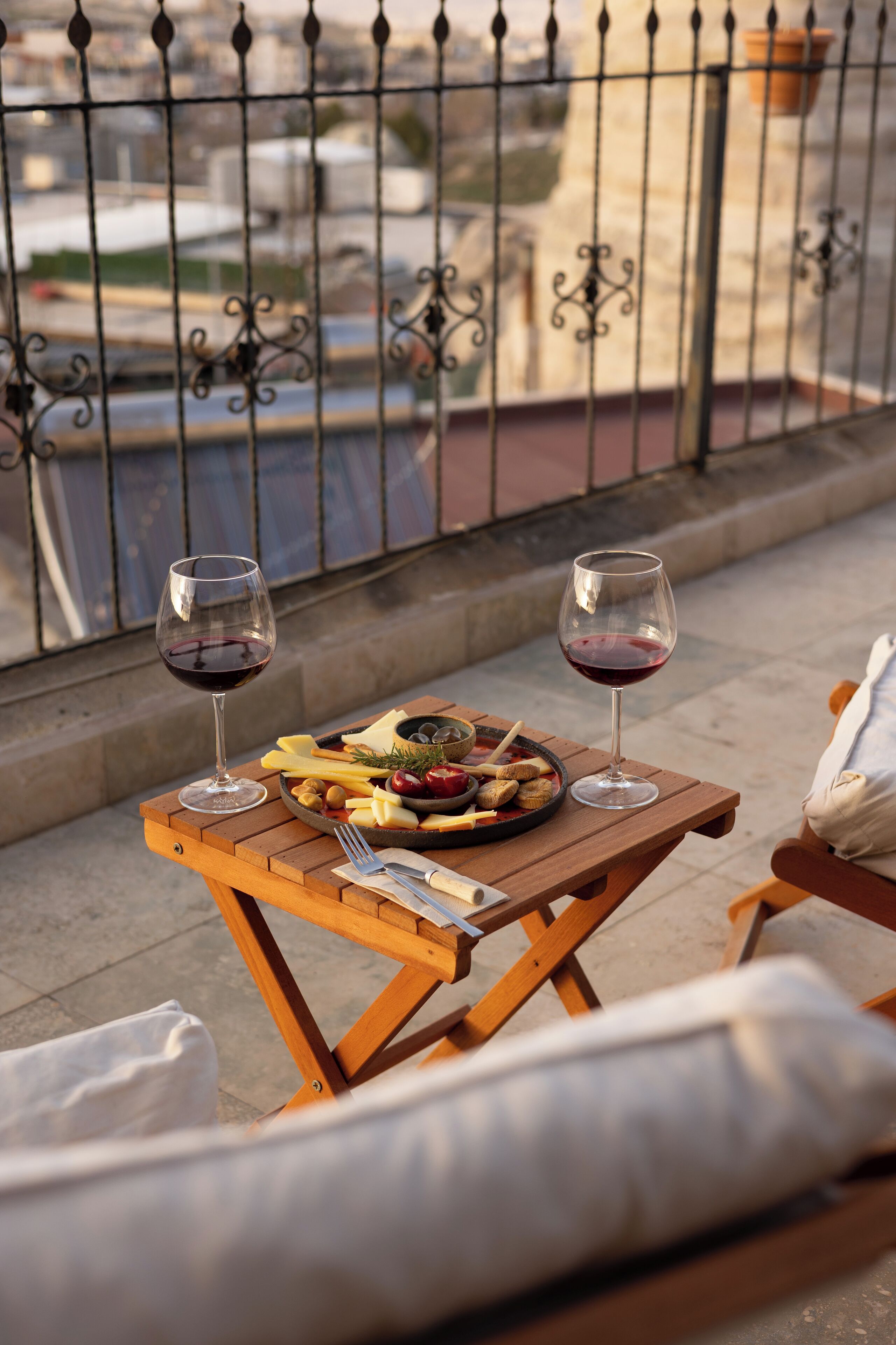 A balcony with a wooden table set for two, featuring a plate of cheese and fruit, two glasses of red wine, and a small bowl of nuts.