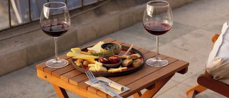 A balcony with a wooden table set for two, featuring a plate of cheese and fruit, two glasses of red wine, and a small bowl of nuts.