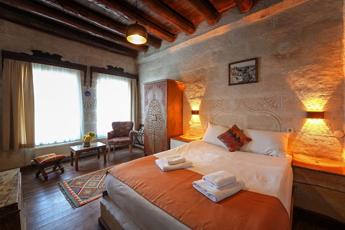 Habitación de hotel con una cama grande, cabecera de madera, una mesita con un florero, una silla y una ventana con cortinas.