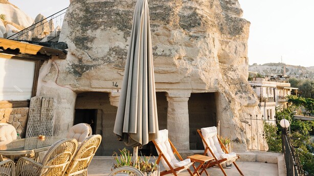 Un edificio de piedra con un exterior singular que se asemeja a una roca, zona de estar al aire libre con sillas de mimbre y una mesa, y vistas del paisaje circundante.