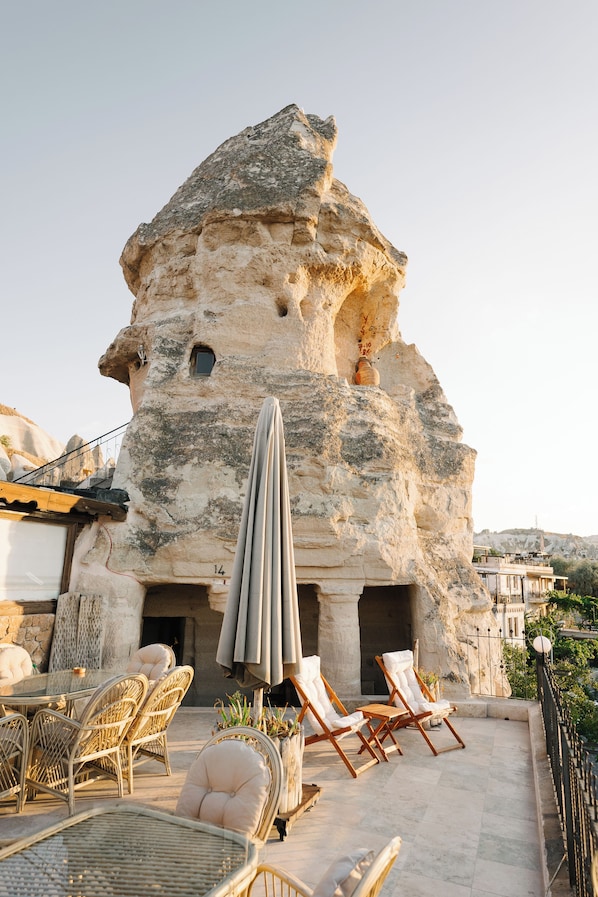 Un edificio de piedra con un exterior singular que se asemeja a una roca, zona de estar al aire libre con sillas de mimbre y una mesa, y vistas del paisaje circundante.