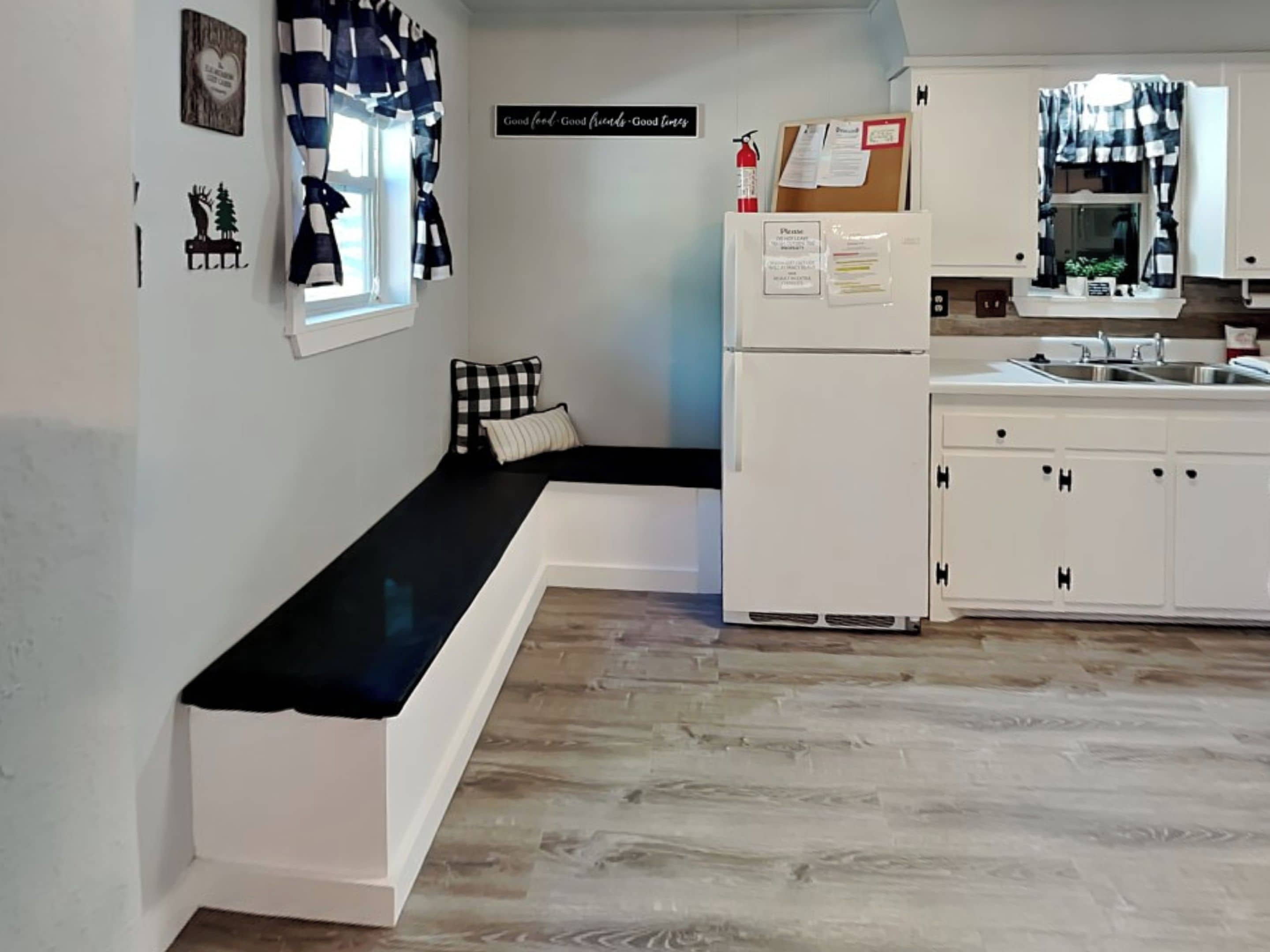 A compact kitchen with a built-in bench, white cabinetry, a window with checkered curtains, and a wall-mounted bulletin board.