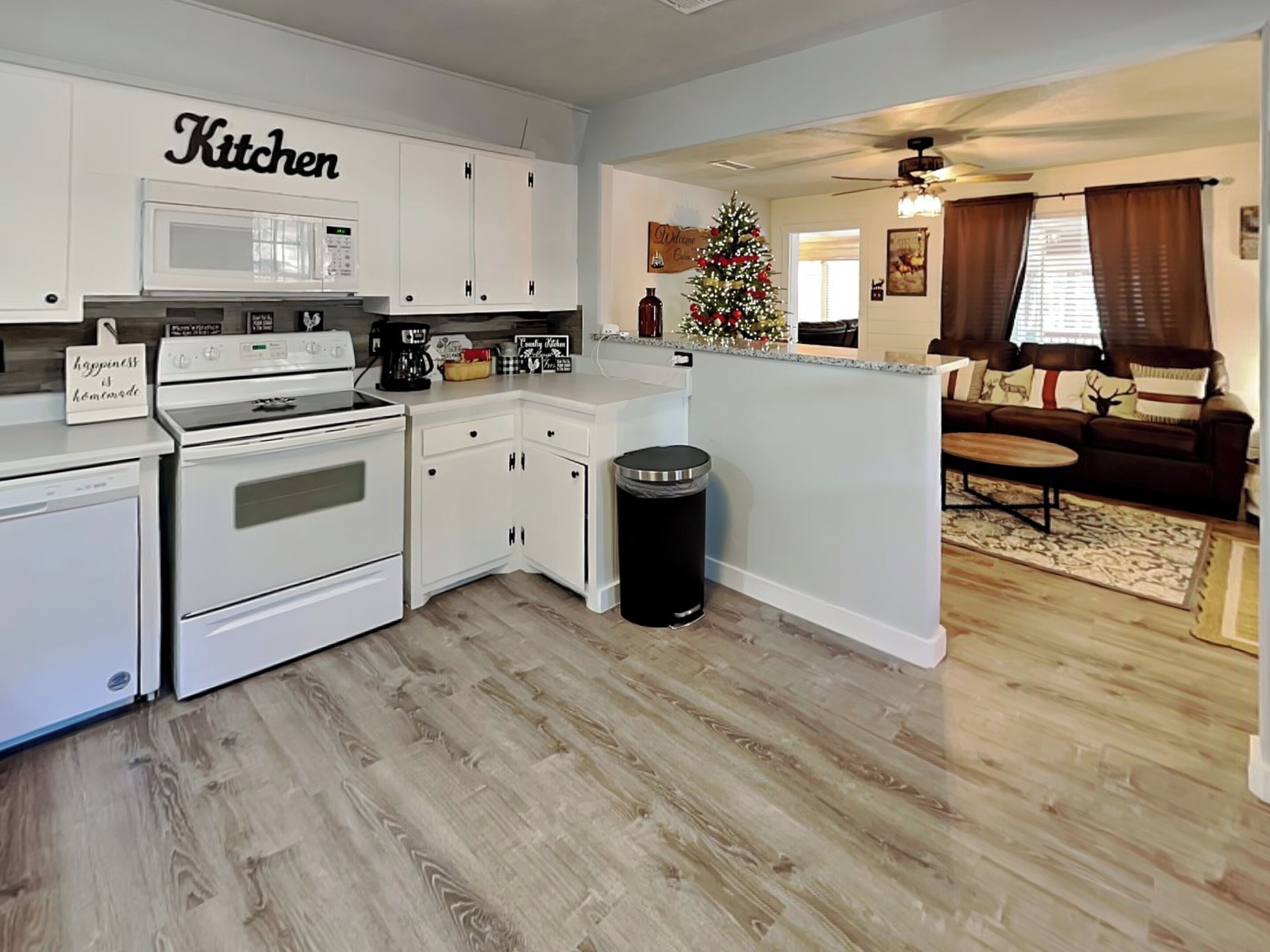 A modern kitchen with white cabinets, a microwave, and a stove. There is a trash can and a Christmas tree in the living area.