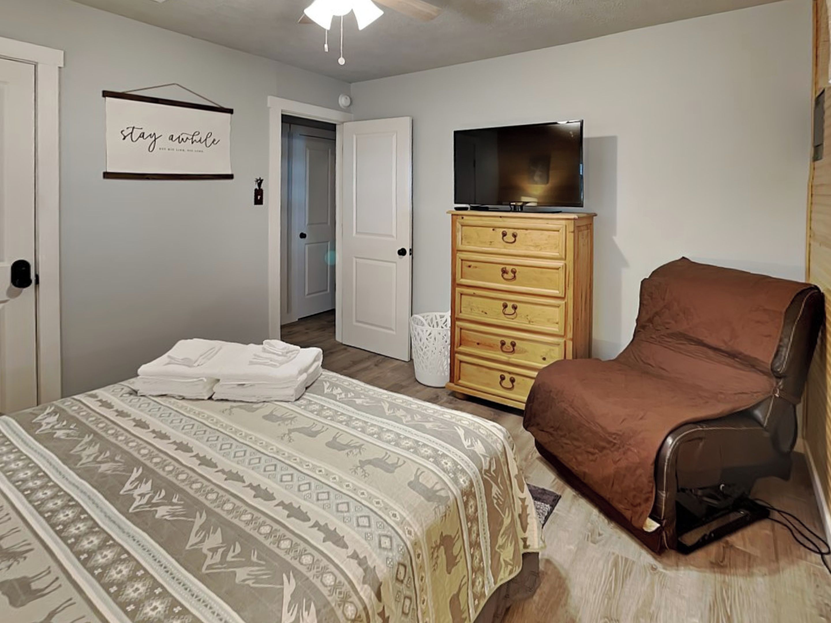 A bedroom with a bed, a dresser, a chair, a television, and a door leading to another room.
