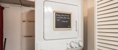 Lavadora blanca con un letrero de pizarra que indica limpiar el filtro de pelusas después de cada ciclo.