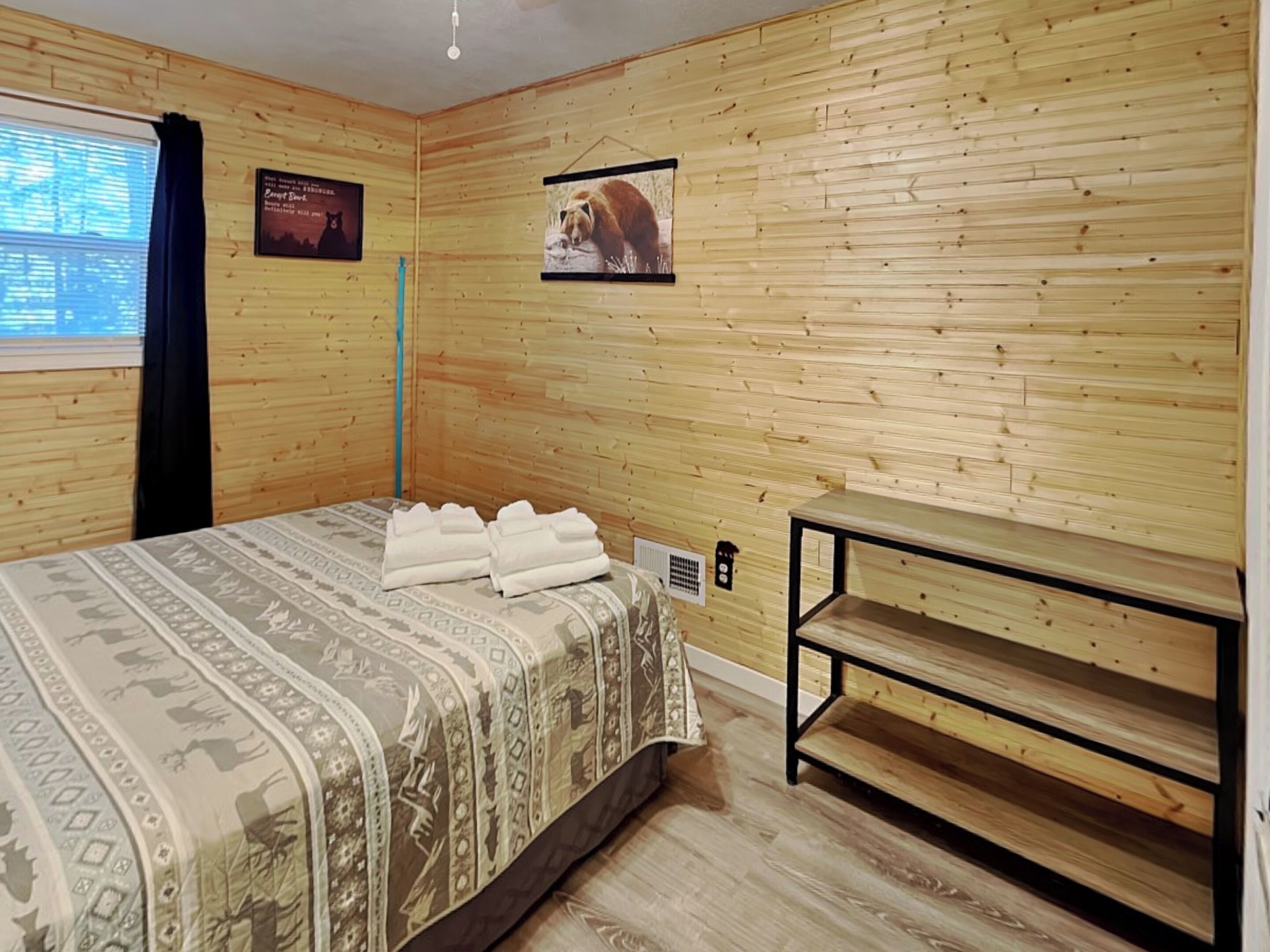 A room with wooden panel walls, a bed with patterned bedding, a small table, and two framed pictures.