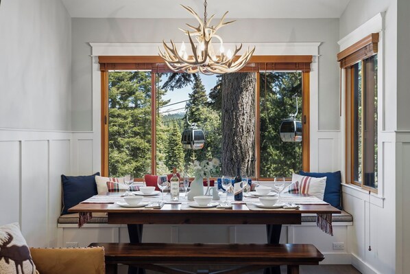 A dining area with a wooden table set for four, a chandelier, and a view of a tree and cable car outside. - 2 Queens | Gondola Views | Rec Center Access | Near Village & Trails (Truckee)