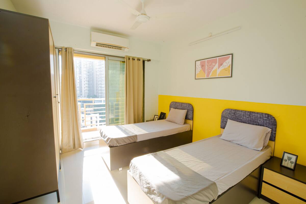 A hotel room with two beds, a ceiling fan, an air conditioning unit, a window with curtains, and a framed picture on the wall.