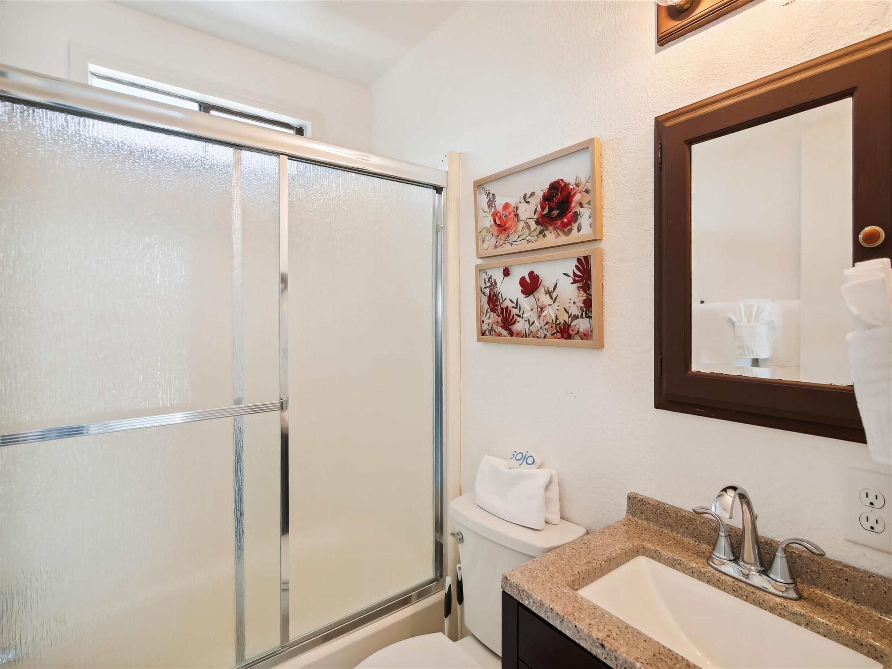 Ein Badezimmer mit Duschkabine aus Glas, Toilette, Waschbecken mit Granitplatte und zwei gerahmten floralen Kunstwerken an der Wand.