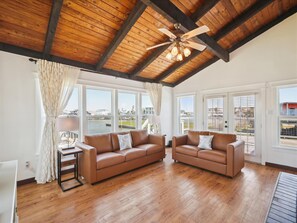 A spacious living room with two brown leather sofas, a wooden ceiling with exposed beams, and large windows offering views of a body of water and buildings. - Tranquil Waters by Ryson Vacation Rentals (Galveston)