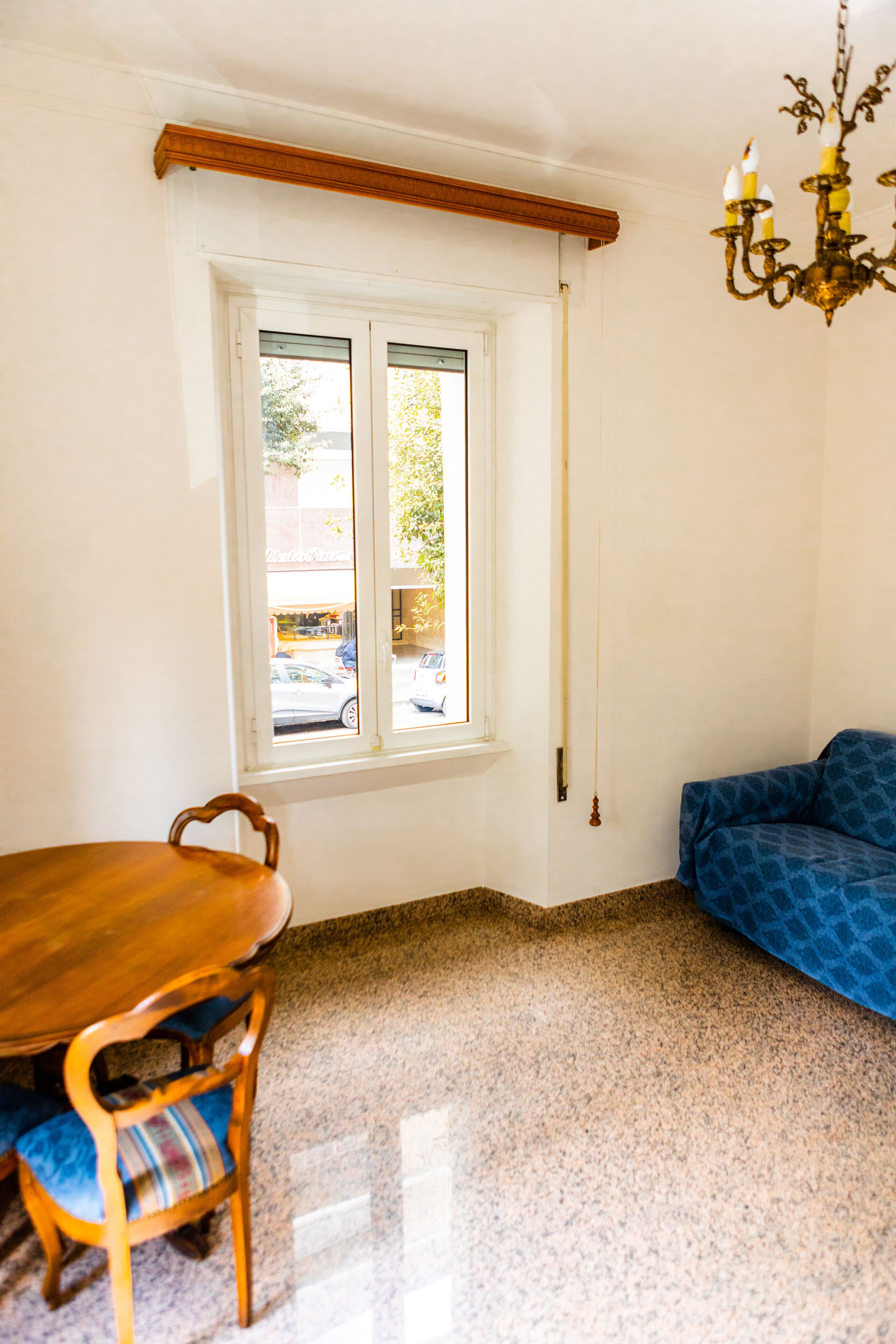 A room with a dining table and chairs, a blue sofa, a window with a view of the street, and a chandelier.