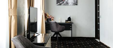 A hotel room with a black and gold patterned carpet, a desk with a computer, a chair, a small table with a vase of flowers, and a painting on the wall.