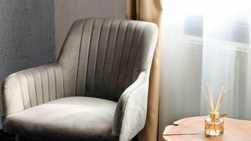 A grey upholstered armchair with vertical ribbing, positioned next to a window with sheer curtains, and a small table with a glass container holding reed diffusers.