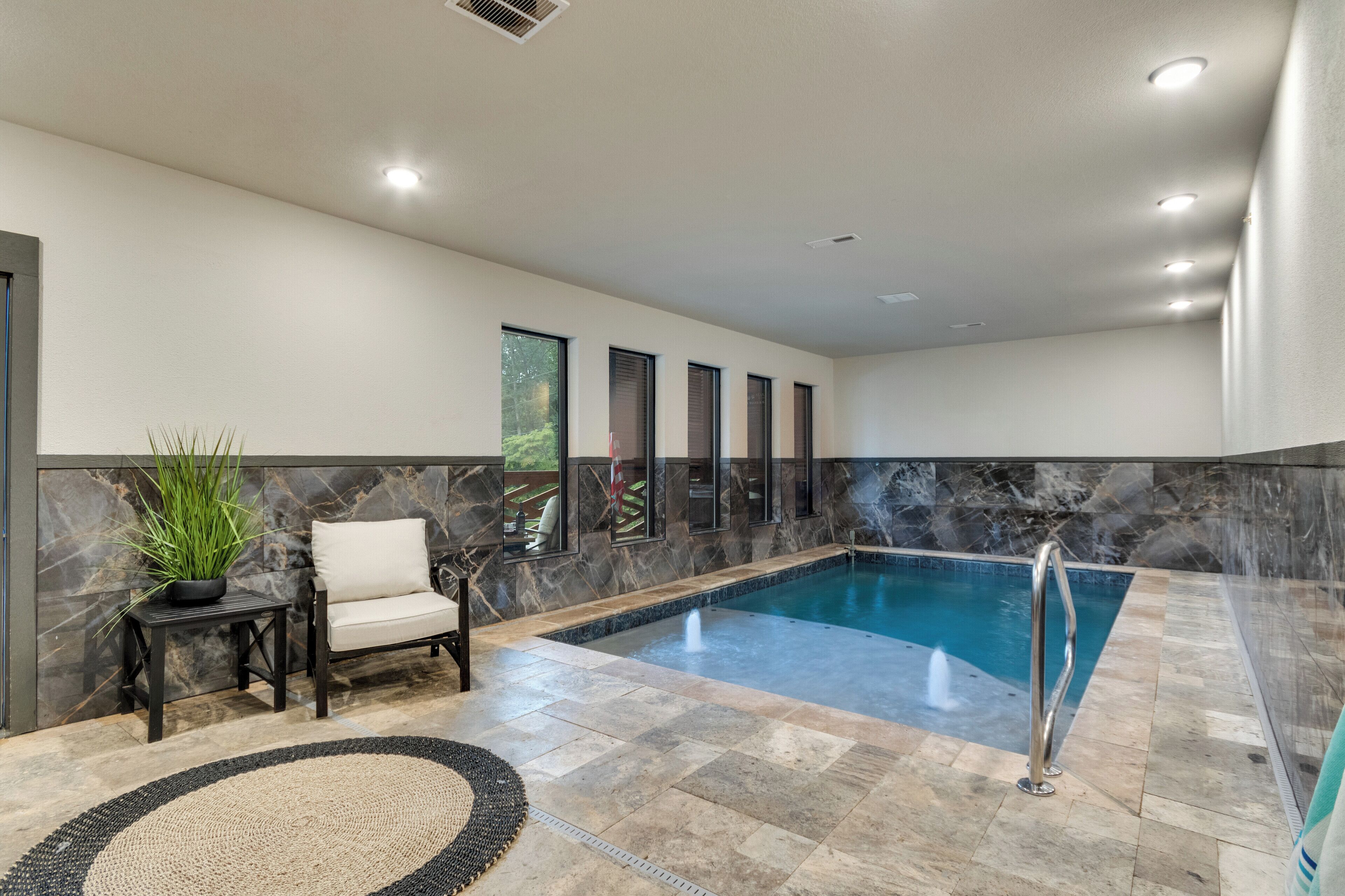 A modern indoor pool area with a marble wall, a white chair, and a small table with a plant.