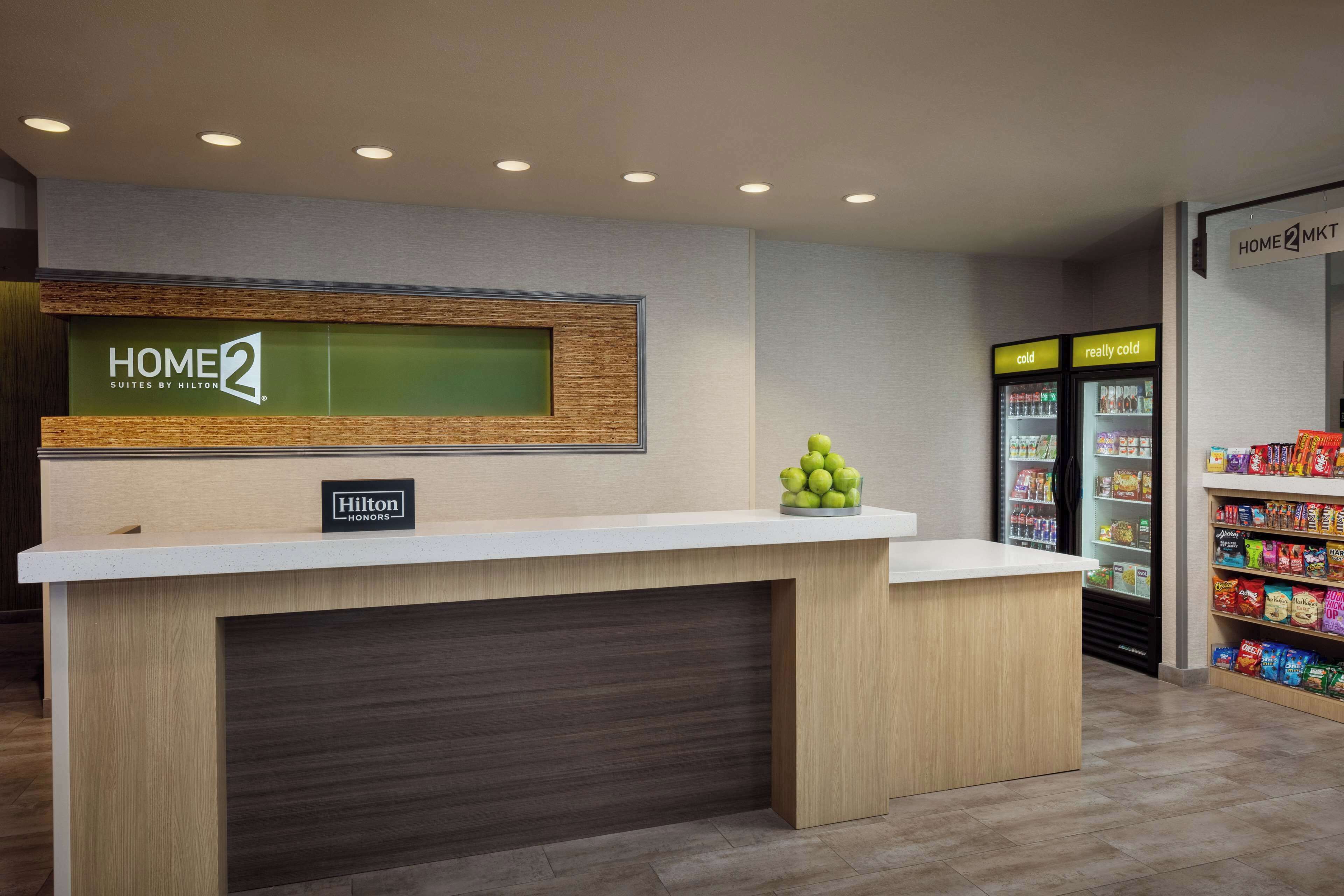 A hotel reception desk with a sign reading "HOME2 Suites by Hilton" and a "Hilton Honors" badge. There are two refrigerators labeled "cold" and "really cold" with snacks and beverages inside.