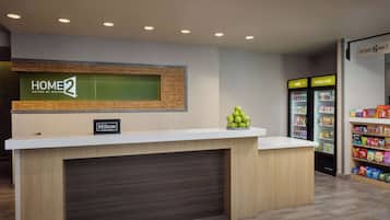A hotel reception desk with a sign reading "HOME2 Suites by Hilton" and a "Hilton Honors" badge. There are two refrigerators labeled "cold" and "really cold" with snacks and beverages inside.