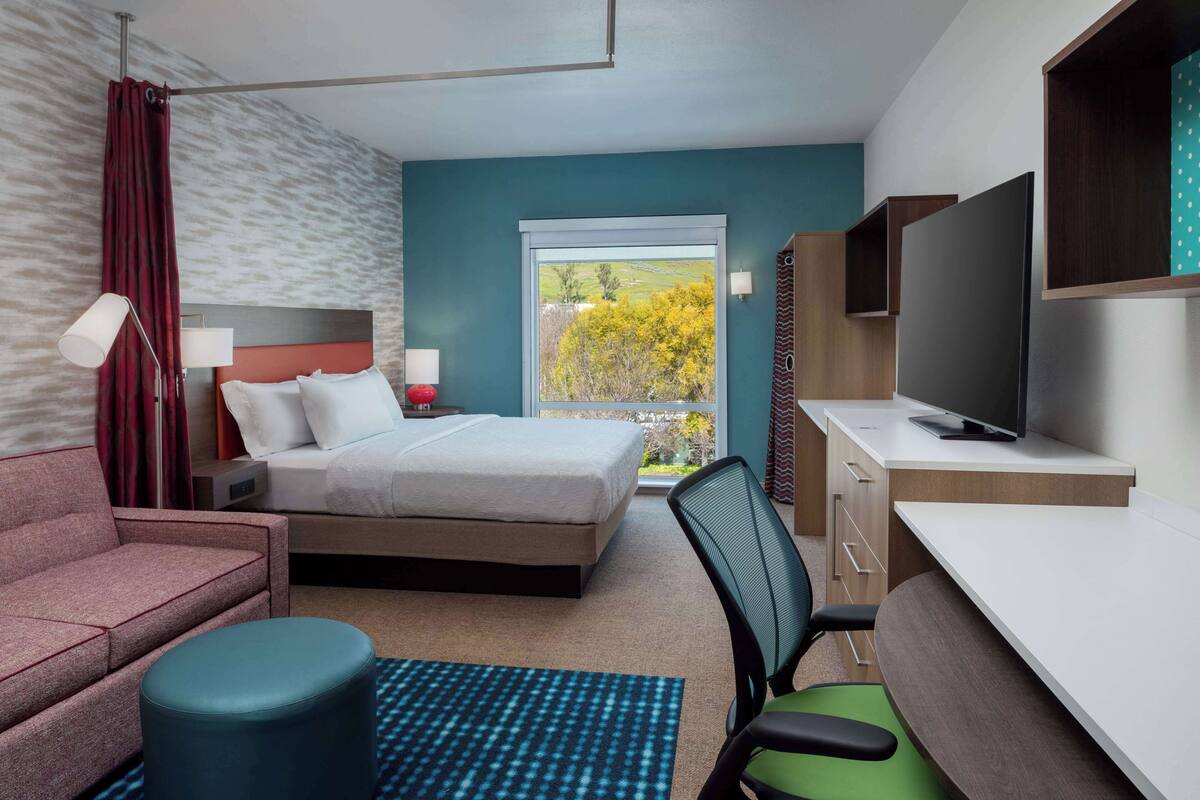 Ein Hotelzimmer mit einem Bett, einem Schreibtisch mit Fernseher, einer Couch, einem Sessel und einem Fenster mit Blick auf Bäume.