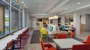 Een moderne eetruimte met lange gedeelde tafels, kleurrijke stoelen en een centrale zithoek met een bank en kussens.