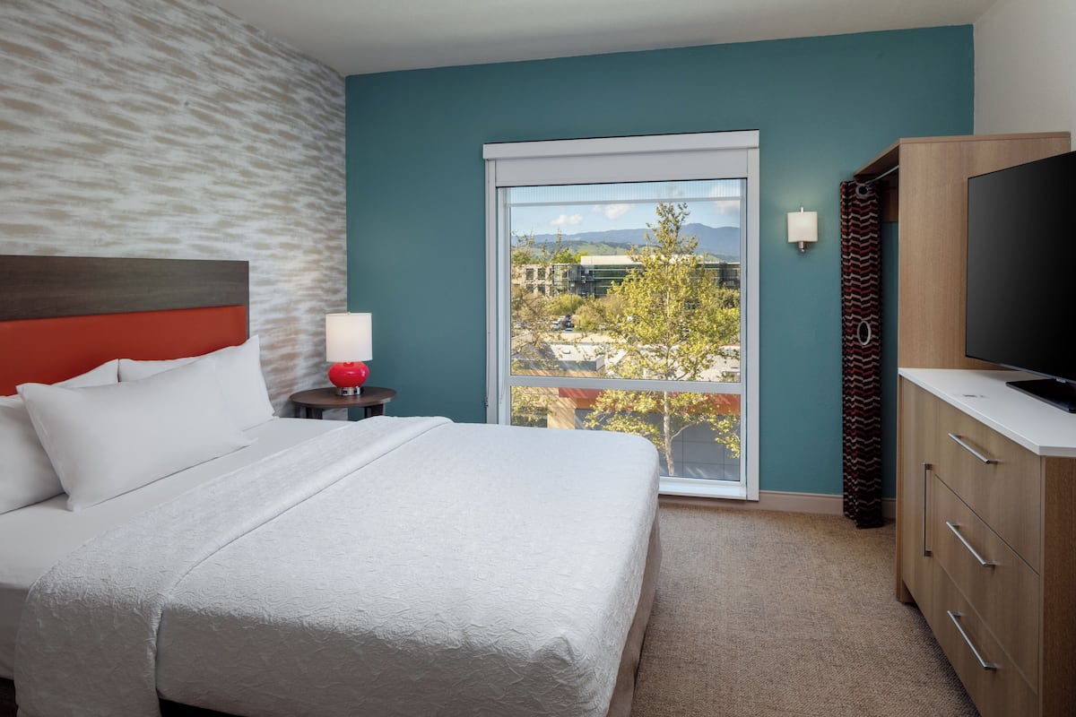Ein Hotelzimmer mit Bett, Fernseher, Nachttisch, Lampe und Blick ins Freie.