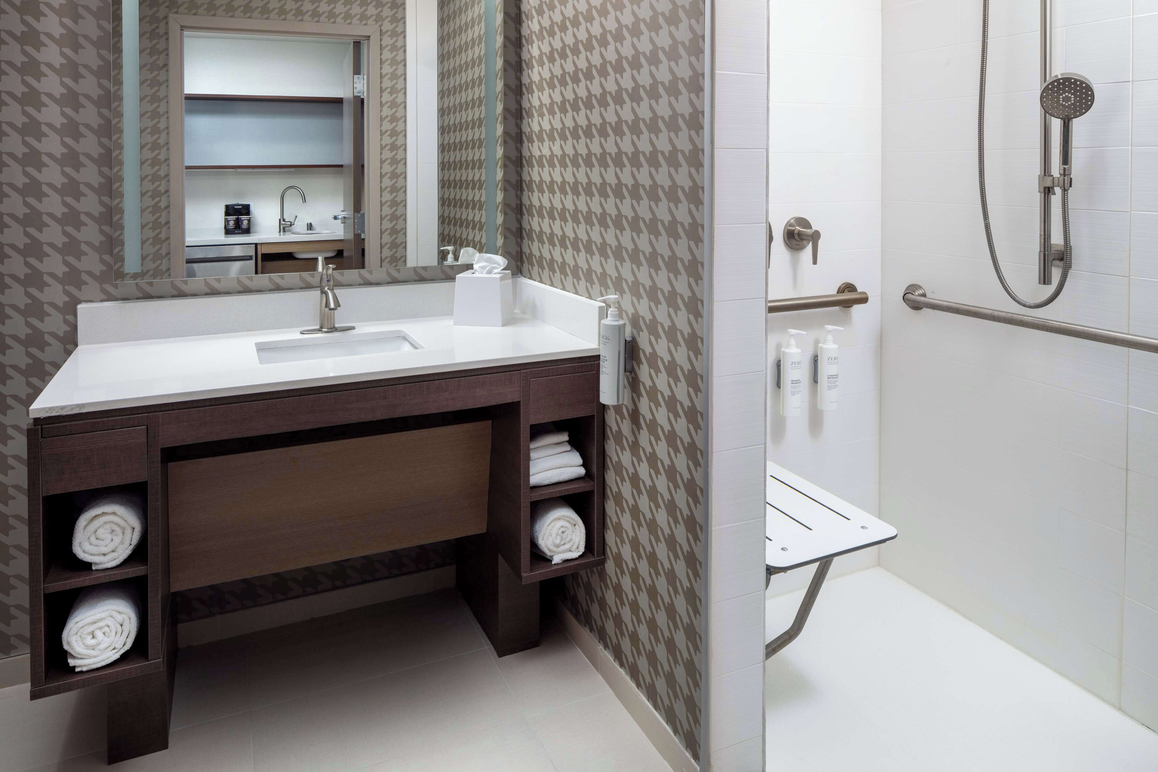 A modern bathroom with a white sink, dark wooden vanity, towel storage, a mirror, and a shower area.