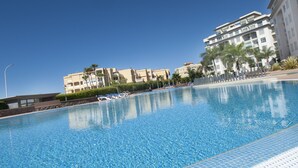 Una piscina con acqua cristallina circondata da una zona con lettini e una palma. Sullo sfondo, edifici a più piani con balconi e un cielo azzurro.