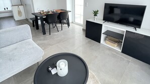 En moderne stue med sofa, rundt sofabord, spiseområde med bord og stole, tv på et sort stativ, samt et stort vindue der lukker naturligt lys ind.