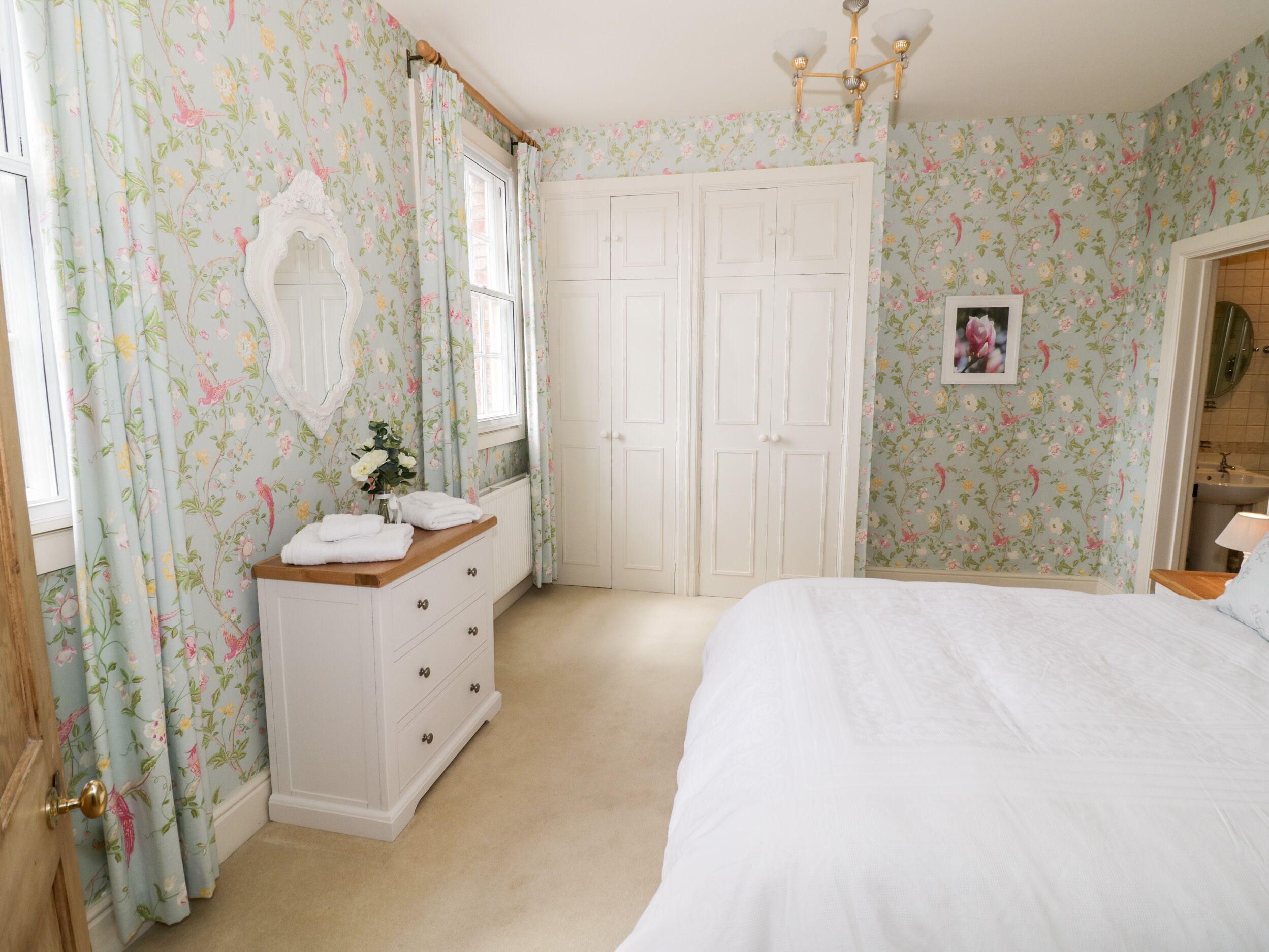Un dormitorio con papel tapiz floral, un tocador blanco, una cama y un armario.