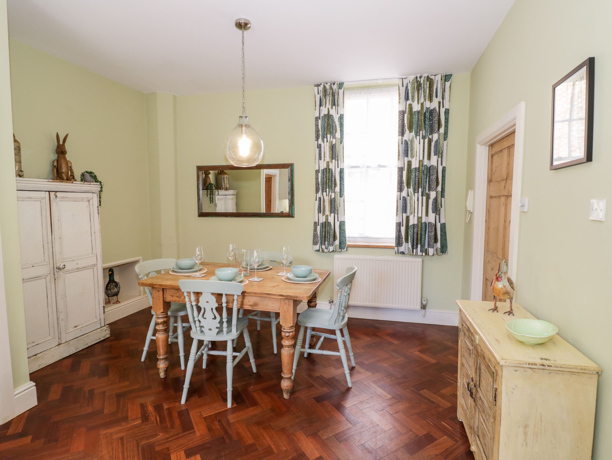 A dining room with a wooden table set for six, surrounded by six chairs. A hanging light fixture is above the table.